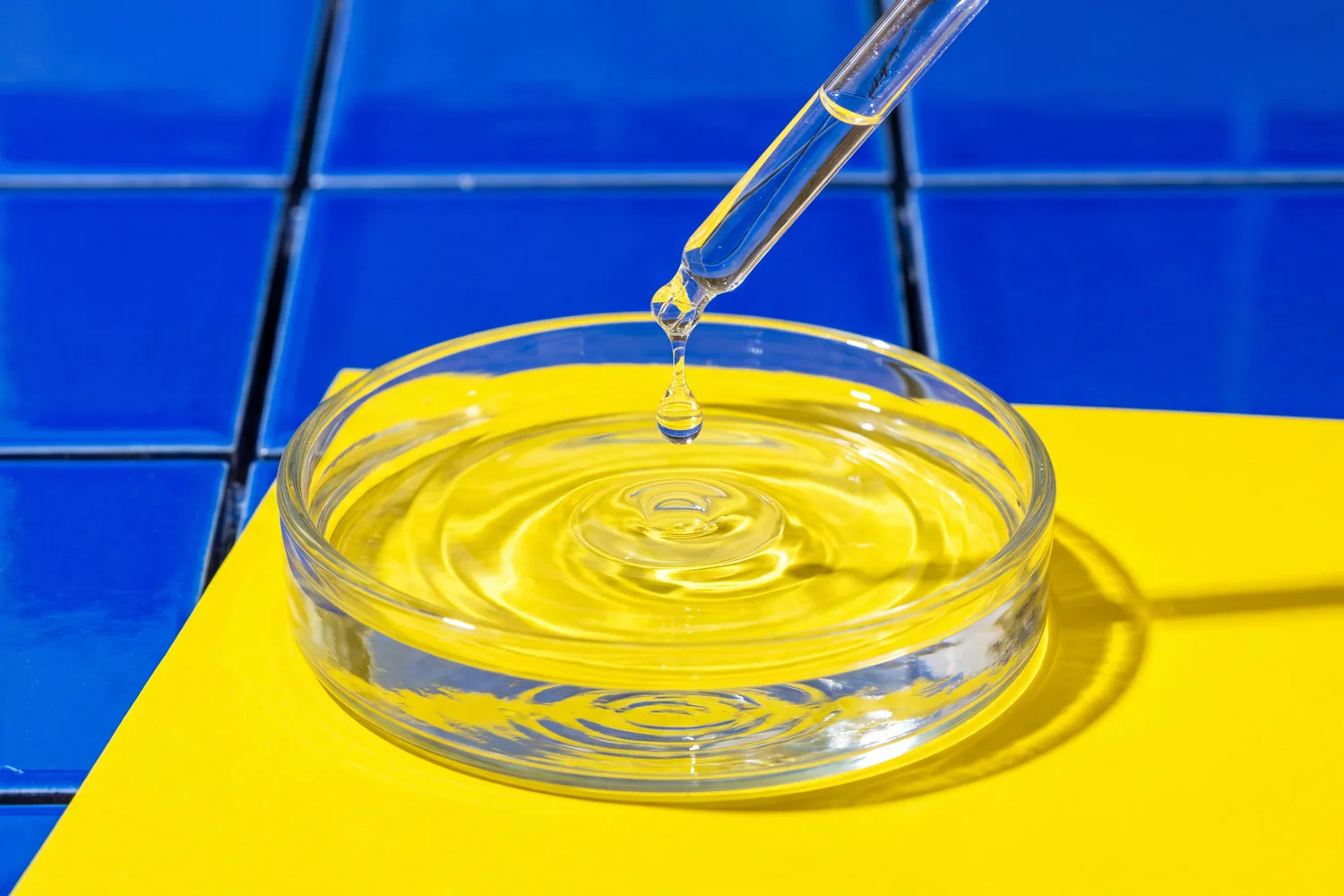 A glass dropper releasing a drop into a clear dish on a yellow counter, cobalt tiles behind.