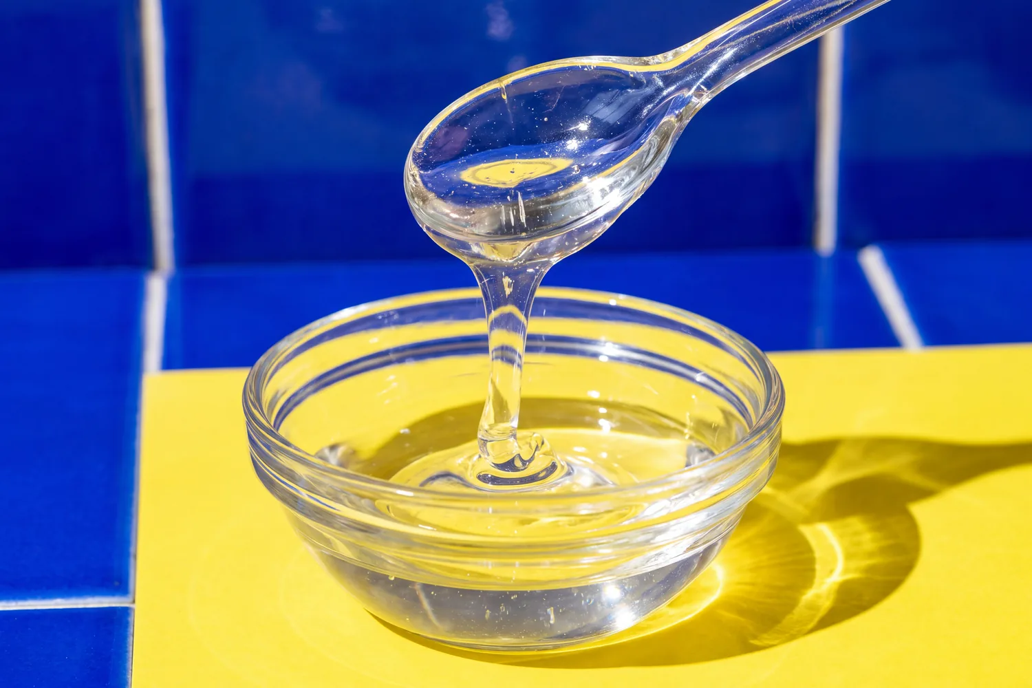 A glass spoon pouring a ribbon of clear glycerin into a glass bowl on a yellow counter, cobalt tiles behind.